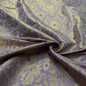 Könnte beinhalten: Nahaufnahme eines luxuriösen Stoffes mit floralem Muster. Der Stoff hat eine weiche Lavendelfarbe mit goldenen Blumenmustern und kleinen lila Akzenten. Der Stoff ist drapiert und zeigt seine Textur und seinen Glanz.
