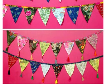 May include: A colorful fabric garland with triangular flags. The flags are made of patchwork fabric with various floral patterns and colors. The garland is strung on a white cord and has tassels hanging from each flag.