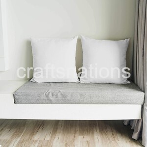 May include: Two white pillows on a gray cushioned bench with a white base. The bench is in front of a white wall and a beige curtain.