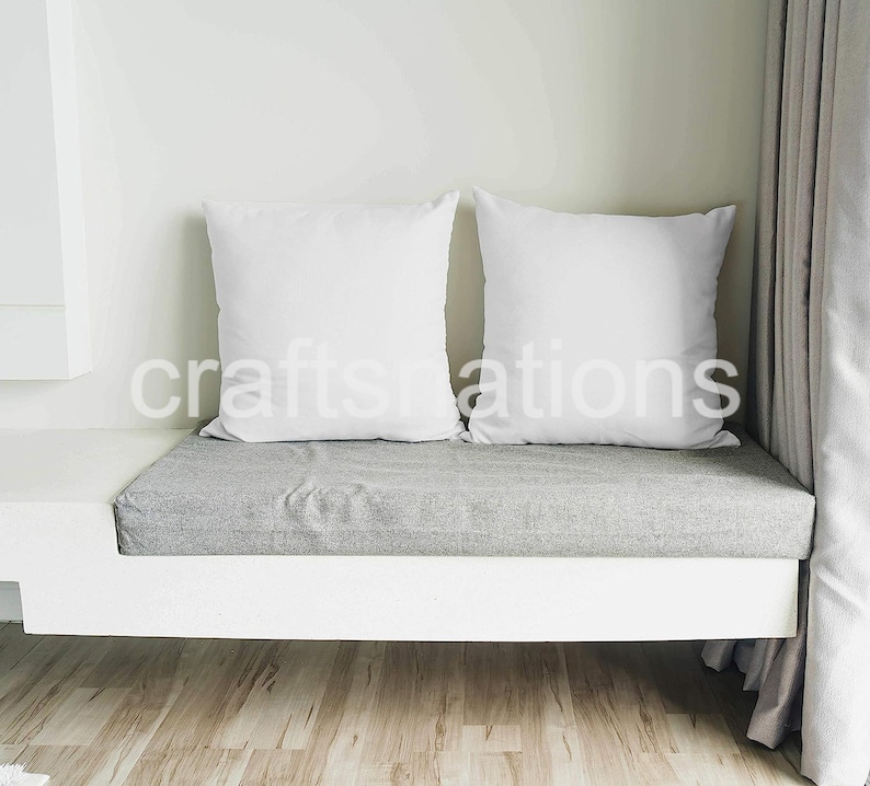 May include: Two white pillows on a gray cushioned bench with a white base. The bench is in front of a white wall and a beige curtain.