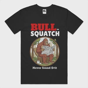 May include: Black t-shirt with the text "BULL SQUATCH" in red and white. The shirt features a cartoon illustration of a Bigfoot reading a newspaper with the headline "Bigfoot Spotter". The text "Never found $#it" is below the illustration.