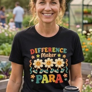 Può includere: Una t-shirt nera a maniche corte con una stampa grafica che riporta il testo "DIFFERENCE Maker PARA" in lettere colorate e testurizzate. Il design include tre margherite bianche con facce sorridenti, braccia e gambe con scarpe color ottanio.