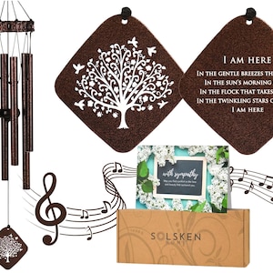 Pode incluir: Um sino de vento com tubos de cor bronze e um design de árvore decorativo. A imagem inclui um cartão de condolências e uma caixa com o texto "SOLSKEN HOME". O sino de vento tem um design de árvore e o texto "I AM HERE".