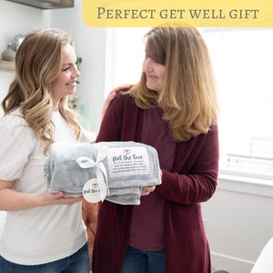 May include: Two people holding a gray fleece blanket with a white tag that says "Feel the Love" and a red heart. The tag has a quote about the power of prayer and the warmth you feel.