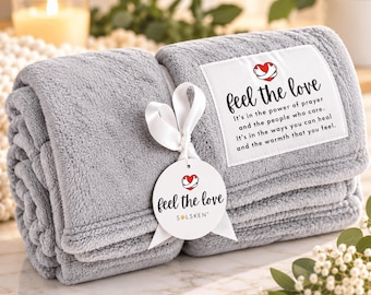 May include: A folded gray plush blanket featuring a white rectangular patch with the text Feel the love, It's in the power of prayer and the people who care. It's in the ways you can heal and the warmth that you feel. A white ribbon holds a round tag.