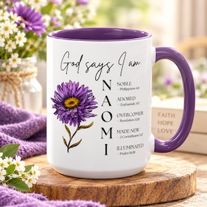 May include: A white ceramic mug with a purple interior and handle, featuring a purple flower illustration and the text God says I am NAOMI followed by the words NOBLE, ADORED, OVERCOMER, MADE NEW, and ILLUMINATED with corresponding Bible verses.