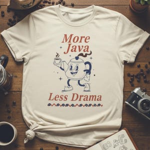 Puede incluir: Camiseta color crema con el texto "More Java Less Drama" en rojo y azul. La camiseta presenta un personaje de tetera de dibujos animados sosteniendo una taza de café. El diseño tiene una estética vintage y retro.