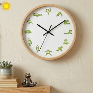 May include: A round wall clock with a light wood frame and a white face featuring twelve illustrations of green frogs in various yoga poses. The clock has three black hands indicating the time, with a clean design and no numbers on the dial.