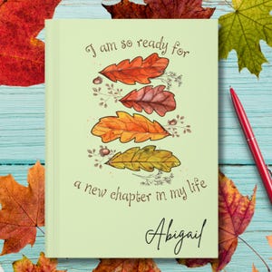 May include: A light green notebook featuring a vertical stack of four autumn leaves in shades of red, orange, and yellow, with the text "I am so ready for a new chapter in my life" and the name "Abigail" printed in a dark brown script font.