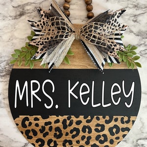 May include: A round wooden sign with a black and leopard print design. The sign reads "MRS. Kelley" in white lettering. It is adorned with a decorative bow featuring animal print and striped ribbons, and green leaf accents.