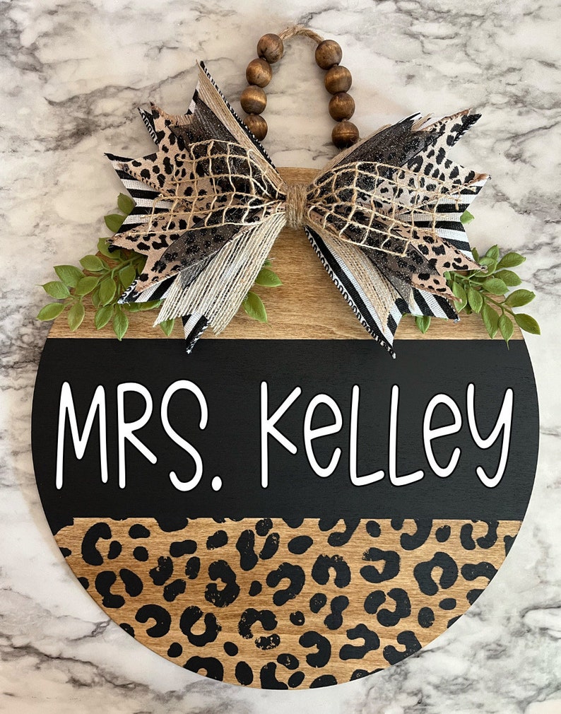 May include: A round wooden sign with a black and leopard print design. The sign reads "MRS. Kelley" in white lettering. It is adorned with a decorative bow featuring animal print and striped ribbons, and green leaf accents.