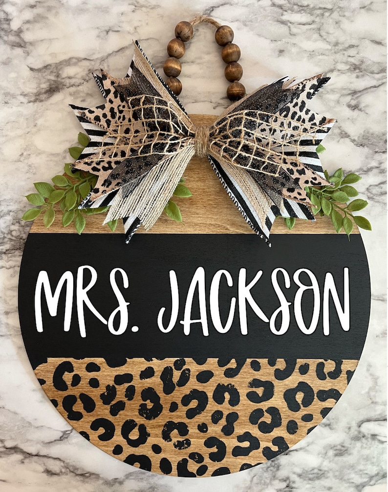 May include: Round wooden sign with a black and leopard print design. The sign features the text "MRS. JACKSON" in white script. A decorative bow with leopard print and greenery accents the top, with a beaded hanger.