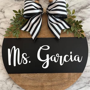 May include: A round wooden sign with a black and white striped bow, green leaves, and the text "Ms. Garcia" in white lettering on a black background.