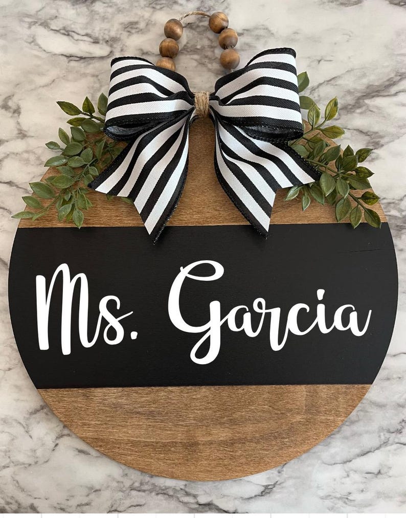 May include: A round wooden sign with a black and white striped bow, green leaves, and the text "Ms. Garcia" in white lettering on a black background.