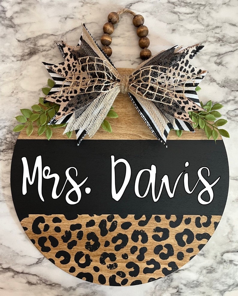 May include: A round wooden sign with a black and leopard print design. The sign has the text "Mrs. Davis" in white script. A decorative bow with leopard print and striped ribbon is attached to the top, along with faux greenery and wooden beads.