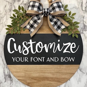 May include: A round wooden door hanger with a black and white checkered bow and greenery. The top half of the hanger is wood and the bottom half is black with white text that says "Customize your font and bow".
