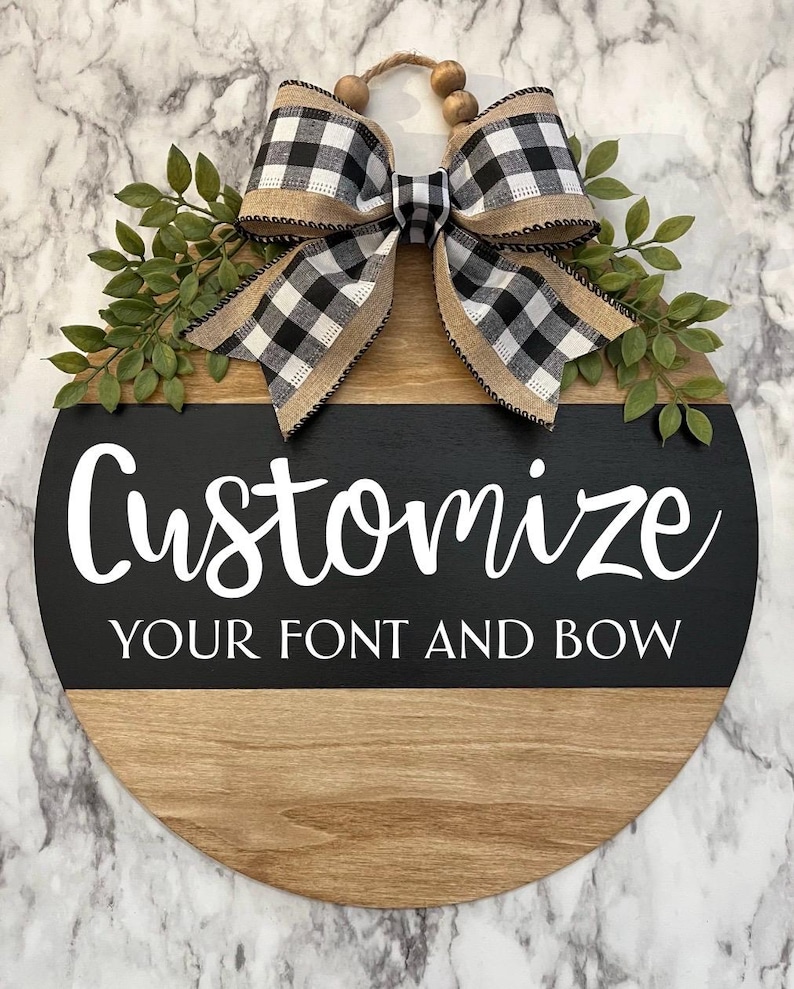 May include: A round wooden door hanger with a black and white checkered bow and greenery. The top half of the hanger is wood and the bottom half is black with white text that says "Customize your font and bow".