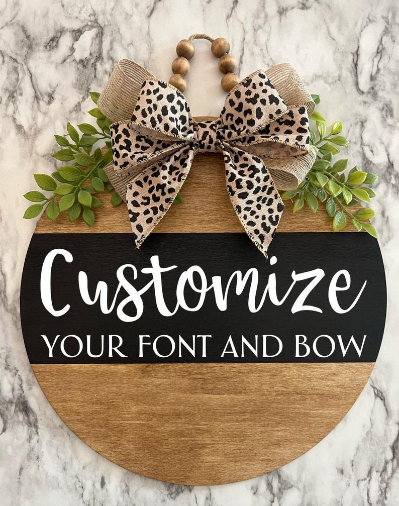May include: A round wooden sign with a black and white stripe across the middle. The sign says "Customize Your Font and Bow" in white text. The sign is decorated with a leopard print bow and green leaves.