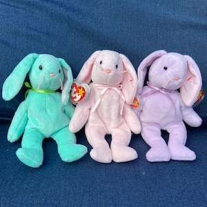 May include: Three plush bunny beanies in different colours: light blue, pink and lavender. Each beanie has a white heart-shaped nose and long floppy ears.