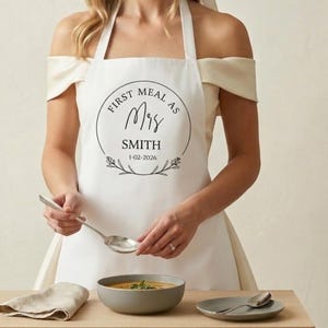 May include: White apron with the text "FIRST MEAL AS Mrs SMITH 1-02-2026" in a black circular design. The apron is being worn by a person. A bowl of soup, a spoon, and a napkin are on a wooden surface.