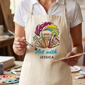 Puede incluir: Delantal beige con un diseño colorido de pinceles y salpicaduras de pintura. El texto "Art with Jessica" está impreso en el delantal. La persona sostiene un pincel y una paleta de madera.