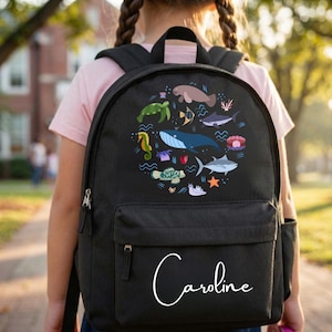 May include: A black backpack featuring a circular graphic of various sea creatures including a whale, turtle, and seahorse. The name Caroline is printed in white cursive script on the front pocket of the bag, which has black straps and zippers.
