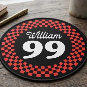 May include: A circular mouse mat featuring a black centre with the white text William and the number 99 in a bold font. The border consists of a red and black chequered pattern. The edges of the mouse mat are stitched with black thread.