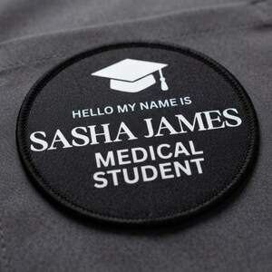 May include: A circular black patch with a white embroidered border and white text. The design features a graduation cap icon above the text HELLO MY NAME IS, SASHA JAMES, and MEDICAL STUDENT, all centred on a dark grey fabric background.