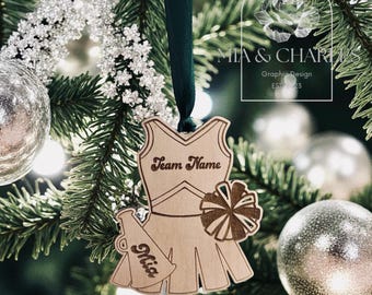 Op de afbeelding: Houten kerstornament in de vorm van een cheerleaderuniform met een groen lint. Het heeft een megafoon en een pompon. De tekst bevat "Team Name" en "Mia". De achtergrond is een kerstboom met zilveren ballen.