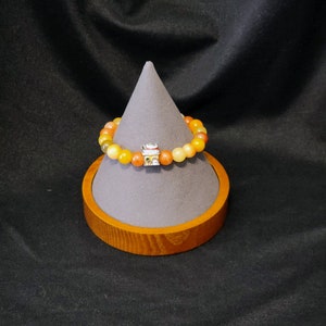 May include: A multi-colored beaded bracelet with a small charm in the center. The bracelet is displayed on a gray and brown stand.