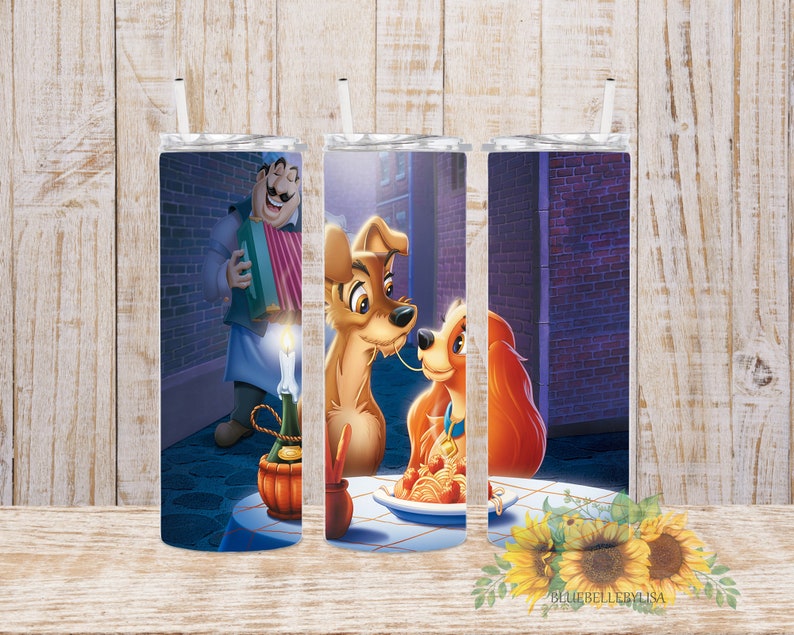 Op de afbeelding: Drie roestvrijstalen bekers met een ontwerp met Lady en de Vagebond die een bord spaghetti delen. De achtergrond is een paarse bakstenen muur met een lantaarnpaal en een man die accordeon speelt.