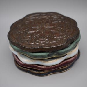 May include: A stack of ceramic coasters in various colors. The top coaster is brown with a floral pattern. Other coasters are green, white, burgundy, and navy blue. The coasters have a wavy edge.