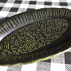 May include: An oval-shaped, dark green ceramic serving platter with a textured surface. The platter features a pattern of yellow-green lines and dashes. The rim has vertical lines, while the center has a cross-hatched design.