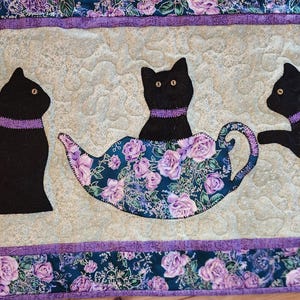 May include: A quilted fabric featuring three black cat silhouettes with purple collars. One cat sits in a teapot with a floral design. The border has a floral pattern with purple accents.