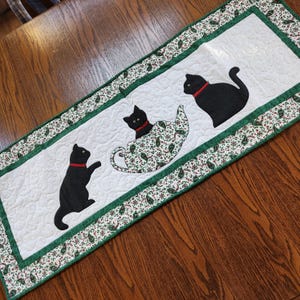 May include: A quilted table runner featuring three black cat silhouettes with red collars. The cats are set against a white background with a paisley teapot design. The border is green with a paisley pattern.