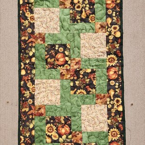 Puede incluir: Un camino de mesa acolchado rectangular con un tema de cosecha de oto&ntilde;o. El camino de mesa presenta un dise&ntilde;o de patchwork con cuadrados de tela verde, crema y negra. La tela negra tiene un patr&oacute;n de girasoles, calabazas y hojas. El camino de mesa est&aacute; bordeado con un borde verde oscuro.