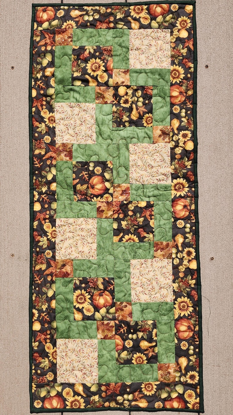 Puede incluir: Un camino de mesa acolchado rectangular con un tema de cosecha de oto&ntilde;o. El camino de mesa presenta un dise&ntilde;o de patchwork con cuadrados de tela verde, crema y negra. La tela negra tiene un patr&oacute;n de girasoles, calabazas y hojas. El camino de mesa est&aacute; bordeado con un borde verde oscuro.