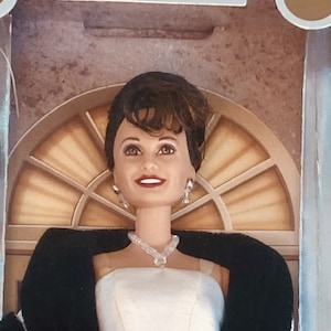 Puede incluir: Muñeca de la serie "Daytime Drama Collection". La muñeca tiene cabello oscuro y lleva un vestido blanco sin tirantes, un chal negro y un collar y pendientes brillantes. El fondo presenta una ventana decorativa.