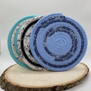 May include: Four round fabric pot holders in blue, black, white, and turquoise. The pot holders are made of fabric scraps sewn together in a spiral pattern. The pot holders are stacked on top of each other on a wooden surface.
