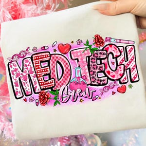 Puede incluir: Una sudadera crema con la frase "MED TECH Crew" en un diseño rosa y rojo. El diseño incluye corazones, flores y gráficos médicos. La sudadera es sostenida por una mano.