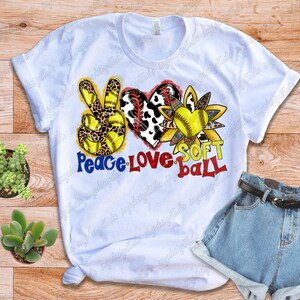 May include: White t-shirt with a peace sign, heart, and sunflower design. The peace sign is yellow and black with leopard print. The heart is red and white with cow print. The sunflower is yellow and brown with a softball in the center. The text "Peace Love Soft Ball" is written below the design.
