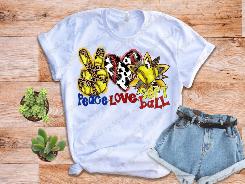 May include: White t-shirt with a peace sign, heart, and sunflower design. The peace sign is yellow and black with leopard print. The heart is red and white with cow print. The sunflower is yellow and brown with a softball in the center. The text "Peace Love Soft Ball" is written below the design.