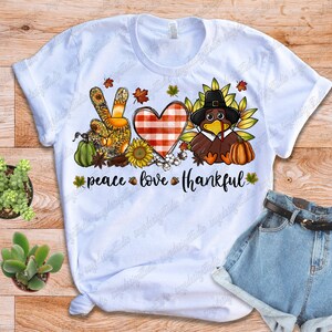 Op de afbeelding: Wit T-shirt met een peace-teken, hart en kalkoen-afbeelding. De afbeelding is in herfstkleuren en bevat de tekst "peace love thankful".