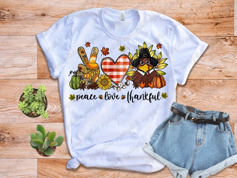 Op de afbeelding: Wit T-shirt met een peace-teken, hart en kalkoen-afbeelding. De afbeelding is in herfstkleuren en bevat de tekst "peace love thankful".
