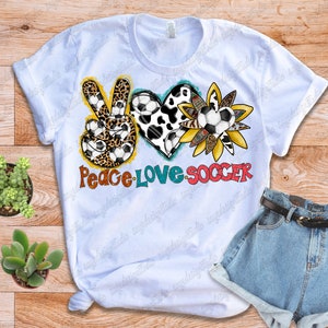 May include: White t-shirt with a peace sign, heart, and sunflower design. The peace sign is made of leopard print and soccer balls. The heart is made of cow print and soccer balls. The sunflower is made of yellow and orange petals and soccer balls. The text "Peace Love Soccer" is written below the design.