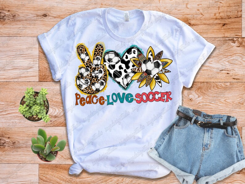 May include: White t-shirt with a peace sign, heart, and sunflower design. The peace sign is made of leopard print and soccer balls. The heart is made of cow print and soccer balls. The sunflower is made of yellow and orange petals and soccer balls. The text "Peace Love Soccer" is written below the design.
