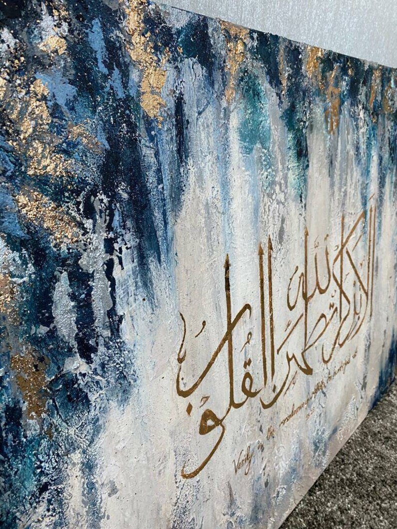 May include: Abstract painting with a blue, white, and gold color scheme. The painting features Arabic calligraphy in gold paint with the text "Verily in the remembrance of Allah"