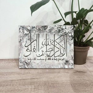 May include: A white canvas with a black and gray textured background. The canvas features a black Arabic calligraphy design with the text "Verily in the remembrance of Allah do hearts find rest" in English below the calligraphy. The canvas is framed and hanging on a wall.