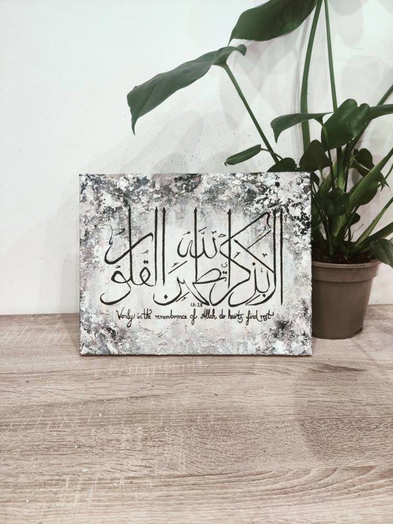 May include: A white canvas with a black and gray textured background. The canvas features a black Arabic calligraphy design with the text "Verily in the remembrance of Allah do hearts find rest" in English below the calligraphy. The canvas is framed and hanging on a wall.
