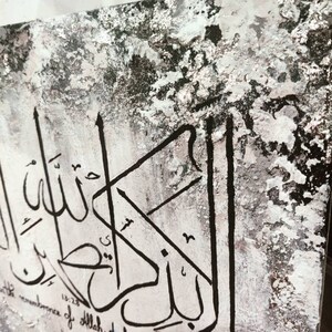 May include: Black and white calligraphy art on a textured canvas background. The Arabic script reads "In the remembrance of Allah do hearts find rest".